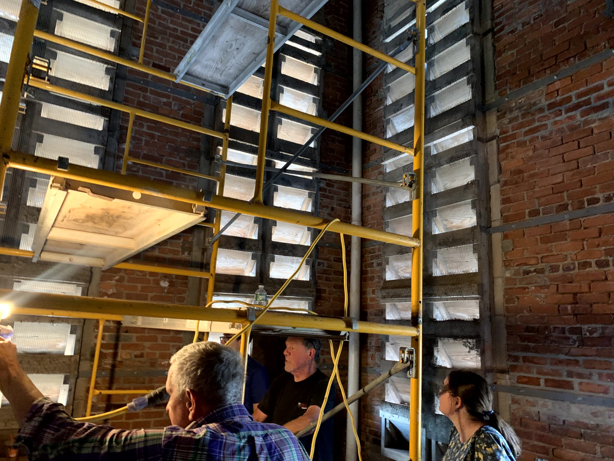 People standing beside interior scaffolding, brick walls, and louvered openings inside the church tower